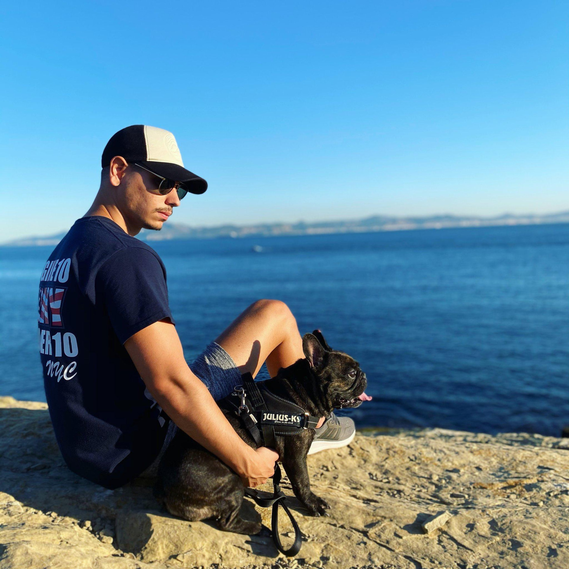 A dog carer sitting with a black French bulldog beside the sea